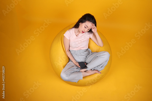 Beautiful young south east Asian woman sits on a yellow beanbag seat orange yellow color background pose fashion style elegant beauty mood expression rest relax think emotion smartphone © oqba