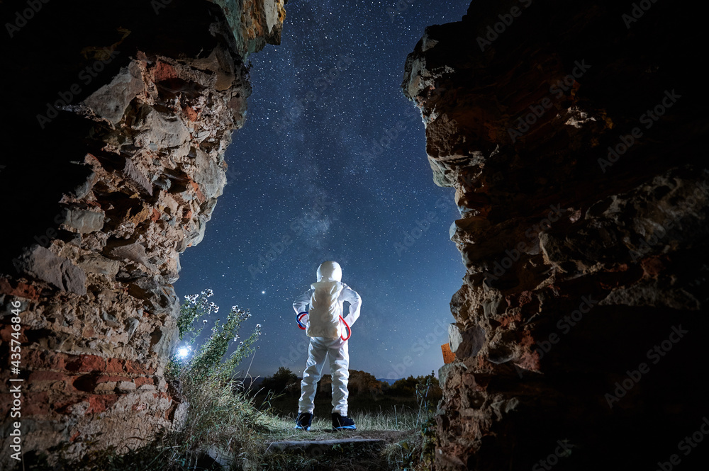 Back view of spaceman in white suit and helmet is standing between abandoned walls at night. Man in spacesuit is looking in starry sky in isolated area. Concept of cosmic traveler.