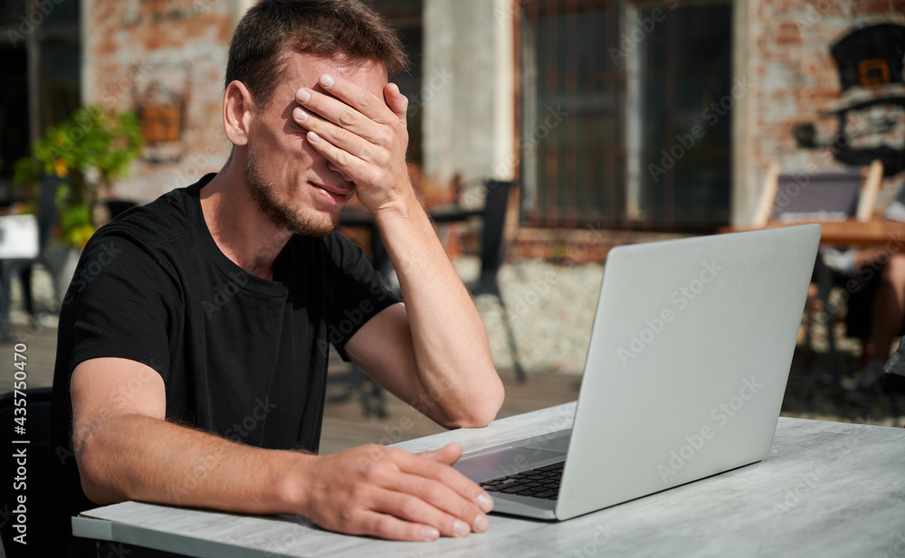 Dissapointed male with hand on face tormented by work. Young man is working on laptop in cafe. Concept of hard worker. Dissapointed male with hand on face tormented by work. Young man is working on laptop in cafe. Concept of hard worker.
