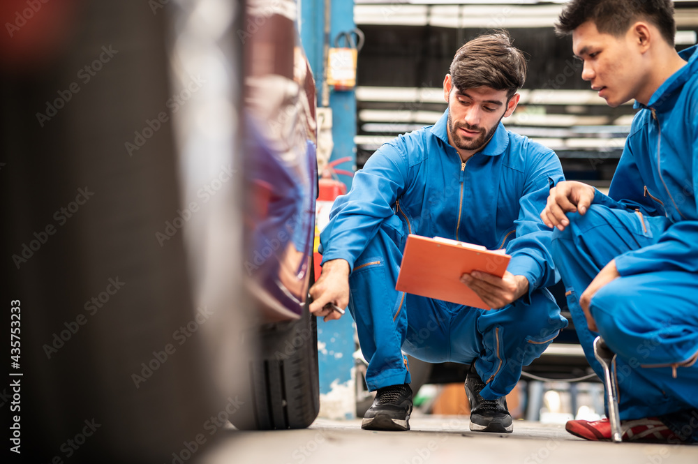 Two professional look technician inspecting car tire and wheel by using check list in moder car service shop. Automotive business or car repair concept.