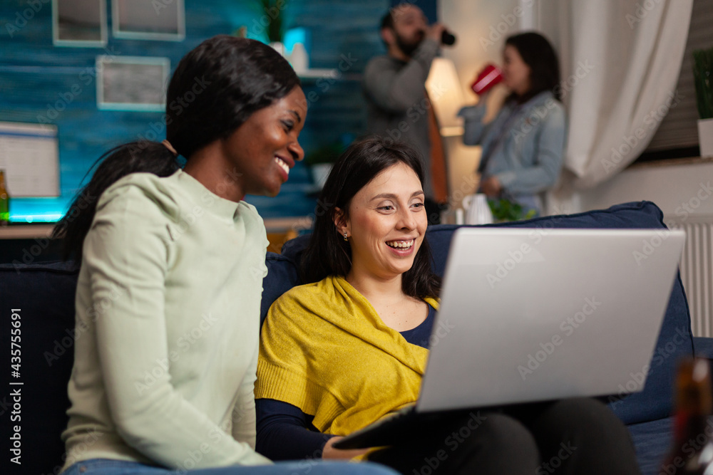 Multi-ethnic friends socializing while watching online funny videos on laptop resting on sofa. In background two womens drinking beer enjoying time spend together during entertainment party. Multi-ethnic friends socializing while watching online funny videos on laptop resting on sofa. In background two womens drinking beer enjoying time spend together during entertainment party.
