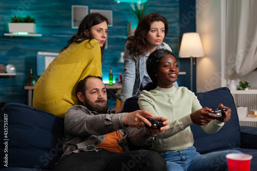 Multi ethnic friends feeling good during oline games challange using wireless controller. Mixed race group of people hanging out together having fun late at night in living room.