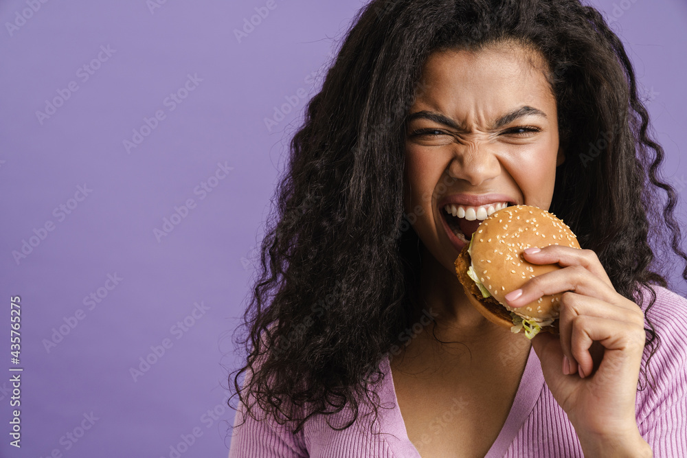 Young black woman with curly hair grimacing while eating hamburger Young black woman with curly hair grimacing while eating hamburger