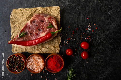Raw piece of ribeye beef meat on on craft paper on a wooden table with tomato, basil, himalayan salt, hot peppers, paprika, spice. Fresh food. Everything for a barbecue.