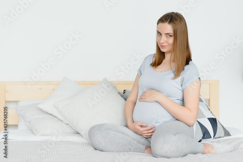 Pregnant young European woman sitting on a large bed and stroking her big belly, 7 month pregnancy, waiting for the birth of her baby.