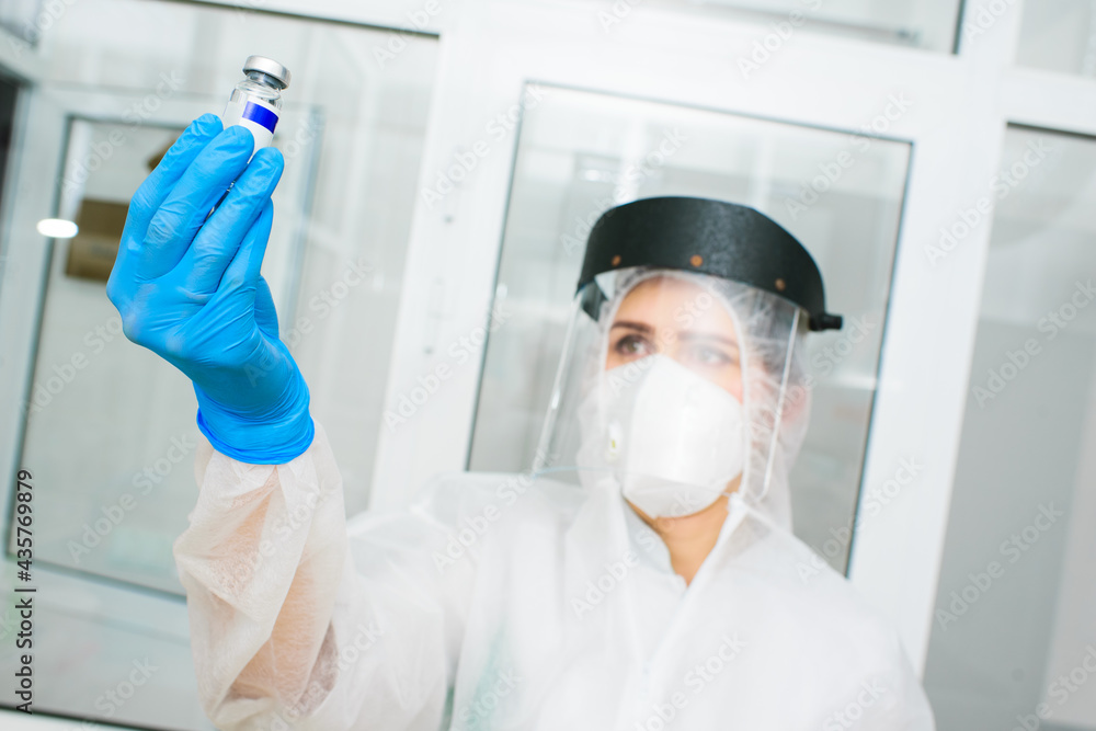 a female doctor or a PCR laboratory researcher in a protective suit, helmet and respirator holds a bottle with a new drug or vaccine in her hands a female doctor or a PCR laboratory researcher in a protective suit, helmet and respirator holds a bottle with a new drug or vaccine in her hands