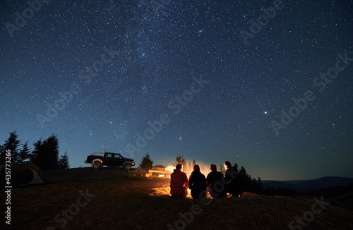 Evening starry sky over mountain valley with car and hikers near campfire. Group of travelers sitting near bonfire under majestic blue sky with stars. Concept of night camping  hiking and travelling.