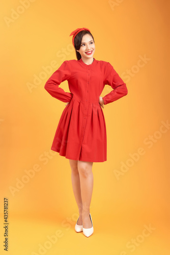 Excited and happy beautiful young woman in red dress is holding hands on hip on yellow background.
