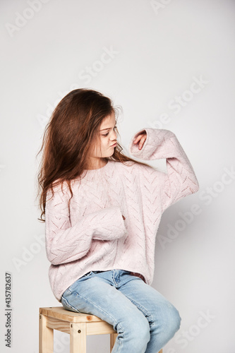 A cute 10-year-old model with an oriental appearance sits on a chair and looks at her hand, dressed in a sweatshirt and jeans, advertising clothes, does not smile. Portrait of a studio photo