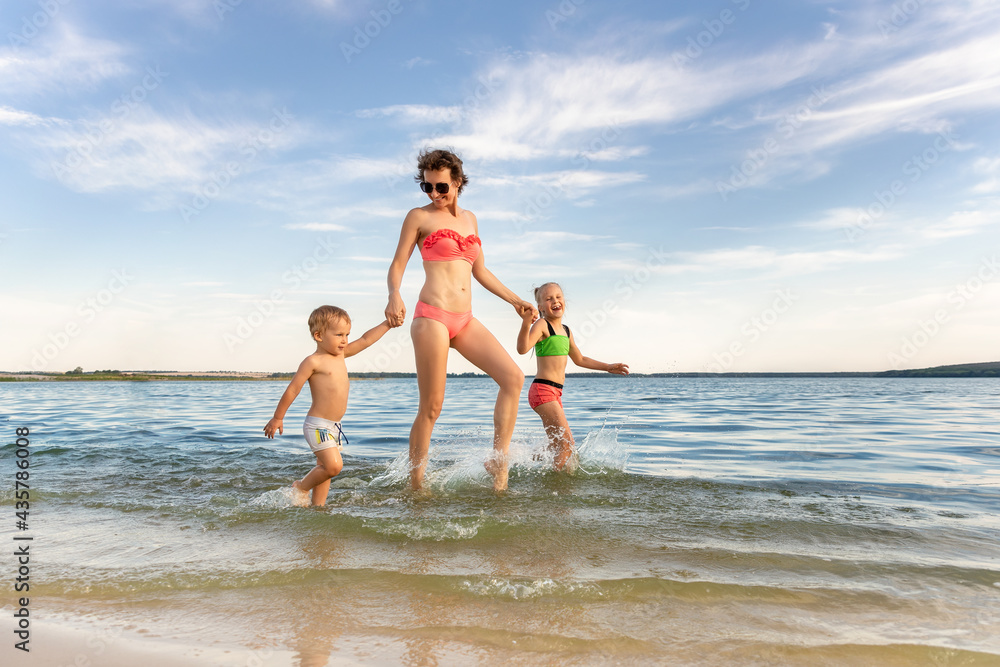 Young adult attractive slim sporty mother enjoy having fun running water by lake or sea sand beach with two cute little siblings against blue sky on summer day. Summertime family vacation concept