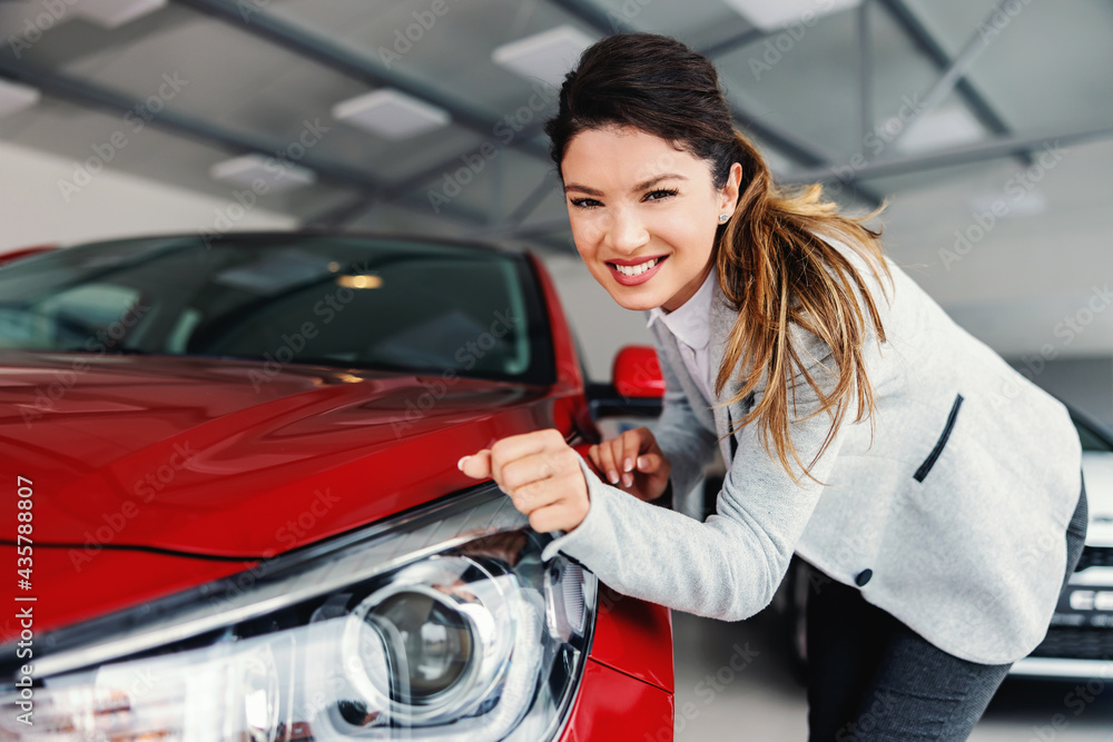 Smiling female car dealer standing in car salon and rubbing headlights with her sleeve while looking at camera. Smiling female car dealer standing in car salon and rubbing headlights with her sleeve while looking at camera.