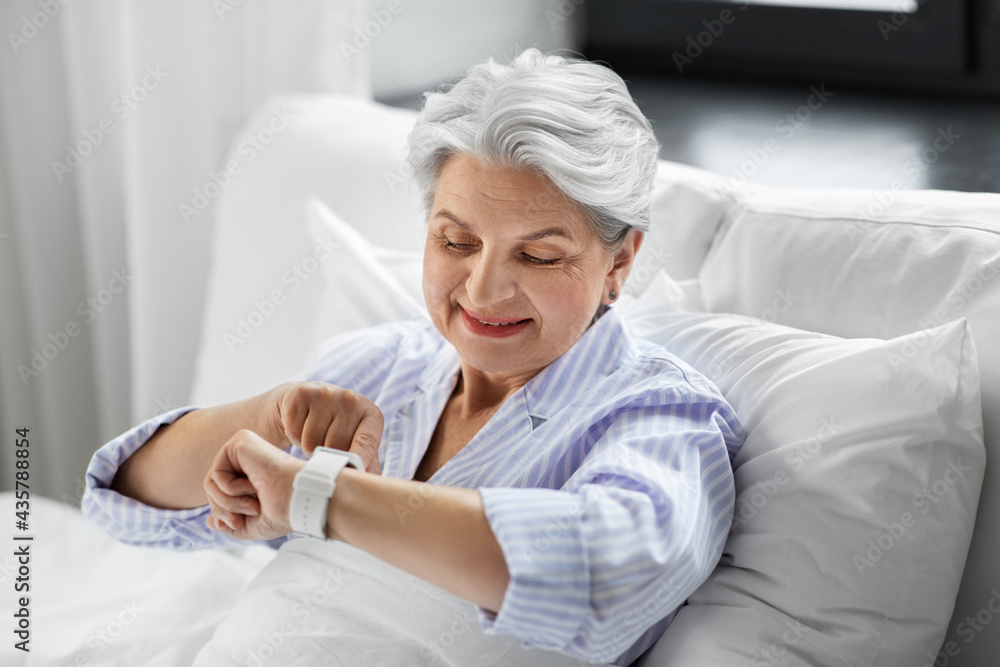 old age and people concept - happy smiling senior woman in pajamas with smart watch sitting in bed at home bedroom old age and people concept - happy smiling senior woman in pajamas with smart watch sitting in bed at home bedroom