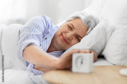 old age and people concept - happy smiling senior woman with alarm clock lying in bed at home bedroom