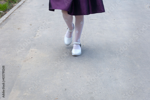 The legs of a girl of six years in beautiful white tights and and elegant white shoes. The child is going on the asphalt
