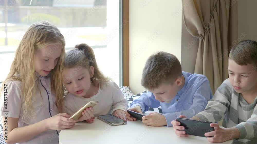 Children sit at a table in a cafe and play mobile phones together. Modern entertainment. A group of friends are sitting at a table in a cafe and everyone is looking at their phone Children sit at a table in a cafe and play mobile phones together. Modern entertainment. A group of friends are sitting at a table in a cafe and everyone is looking at their phone