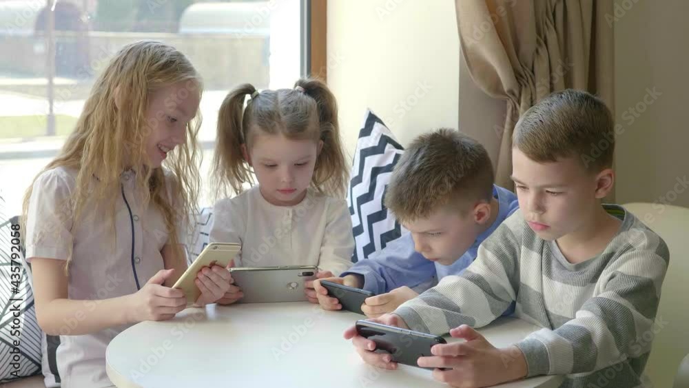 Children sit at a table in a cafe and play mobile phones together. Modern entertainment. A group of friends are sitting at a table in a cafe and everyone is looking at their phone Children sit at a table in a cafe and play mobile phones together. Modern entertainment. A group of friends are sitting at a table in a cafe and everyone is looking at their phone