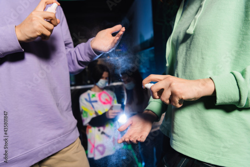 multiethnic friends applying hand sanitizer near blurred gamers in medical masks