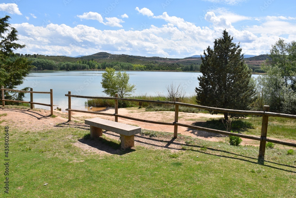 View of La Grajera park from a recreation area. Bench, wooden fence, pine trees and the lake.