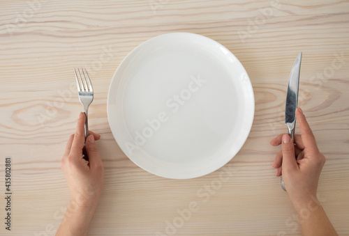 White empty plate and female hands holding silver fork and knife on wooden table background, top view photo