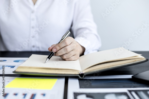 A woman is writing a memo in her notebook, she is reviewing the company financial information and writing unusual notes on the documents the finance department has prepared to bring to the meeting.