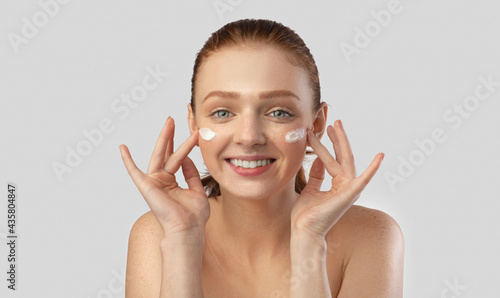 Red-Haired Young Lady Applying Moisturizing Cream On Face, Gray Background