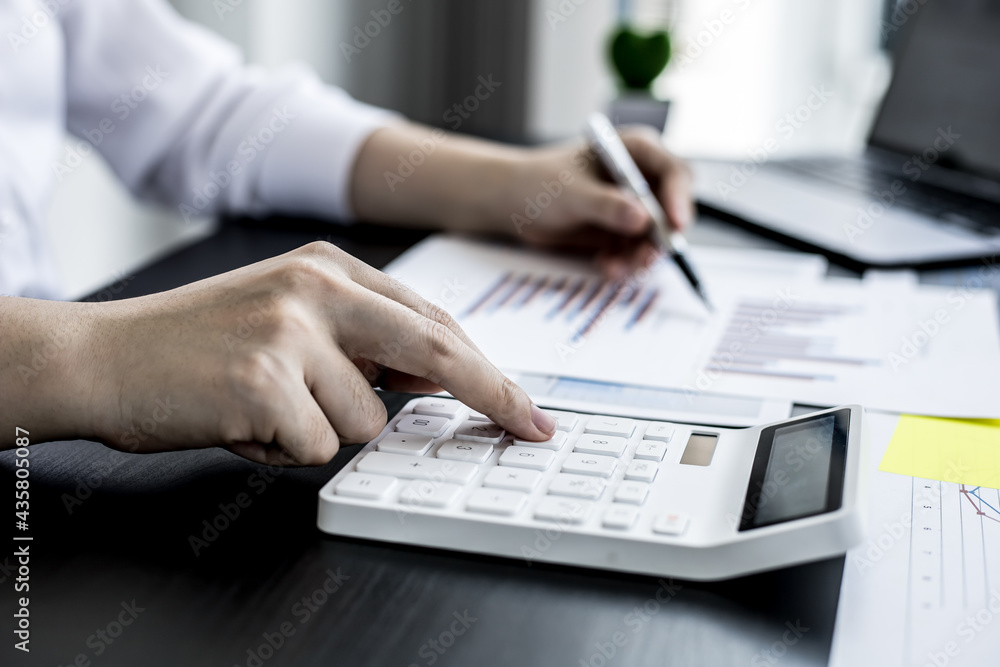 Close-up of woman holding a pen and pressing a white calculator, a corporate finance auditor examining the numbers on financial documents prepared by the finance department to bring to the meeting. Close-up of woman holding a pen and pressing a white calculator, a corporate finance auditor examining the numbers on financial documents prepared by the finance department to bring to the meeting.