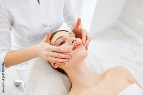 Therapist doing myofascial or buccal massage on face and head for female client lying at beauty center or spa salon
