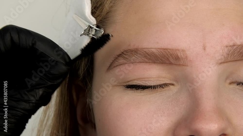 Gloved master shaping eyebrow with a cosmetic brush and pluck eyebrows with tweezers close-up. Stylist's hands combing patient’s eyebrows with a special brush before plucking.