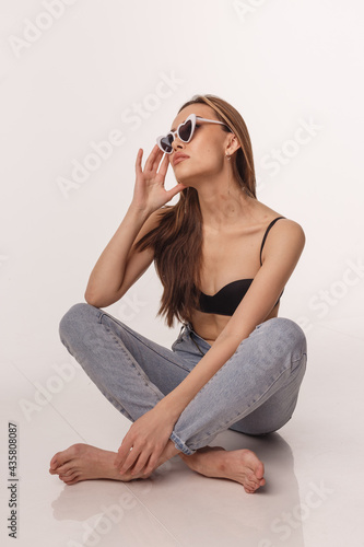 portrait of sexy asian woman with long hair posing in black lingerie, jeans, sunglasses on white studio background with bare feet. model tests of skinny lady in bra. attractive female sitting on floor