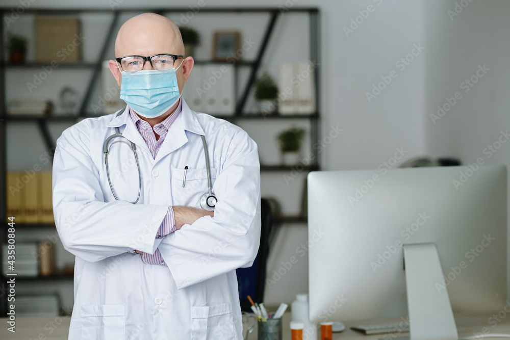 Portrait of confident experienced mature Caucasian doctor in eyeglasses and mask standing with crossed arms in modern office Portrait of confident experienced mature Caucasian doctor in eyeglasses and mask standing with crossed arms in modern office