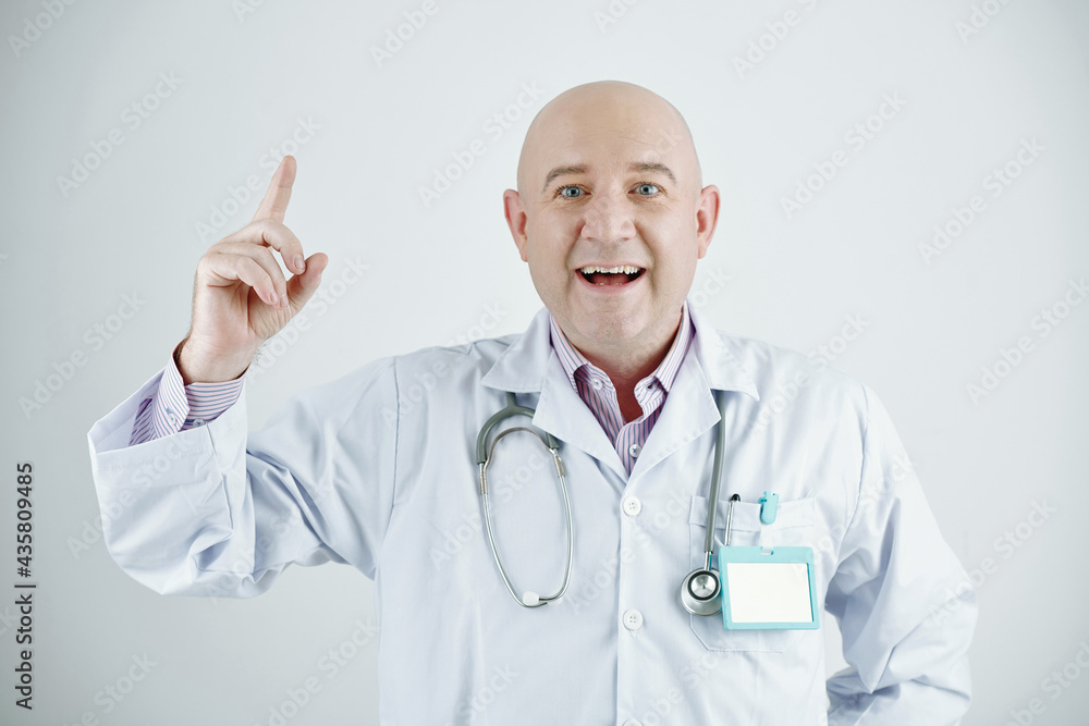 Portrait of cheerful enterprising mature Caucasian doctor in lab coat wearing stethoscope and badge rising up finger while having idea Portrait of cheerful enterprising mature Caucasian doctor in lab coat wearing stethoscope and badge rising up finger while having idea