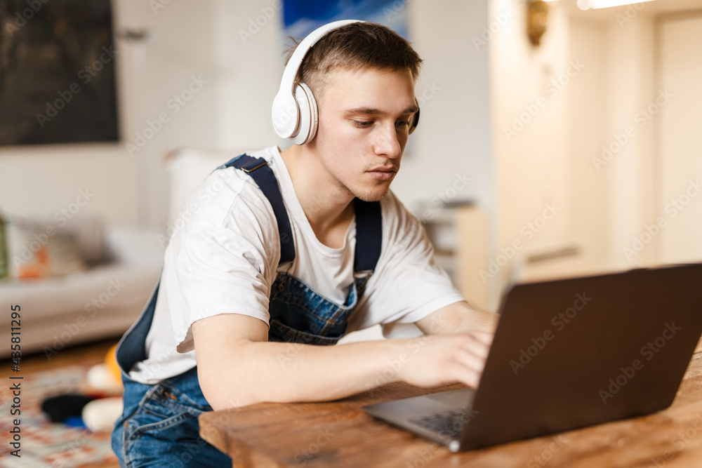 Young man in headphones using laptop while working on craft rug Young man in headphones using laptop while working on craft rug