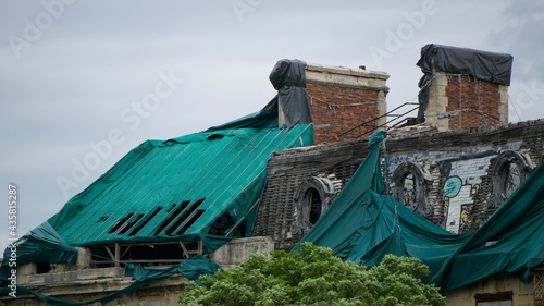 Side view of a dilapidated roof of an old building with two chimneys, graffiti on the walls and some tarp laid out as protection