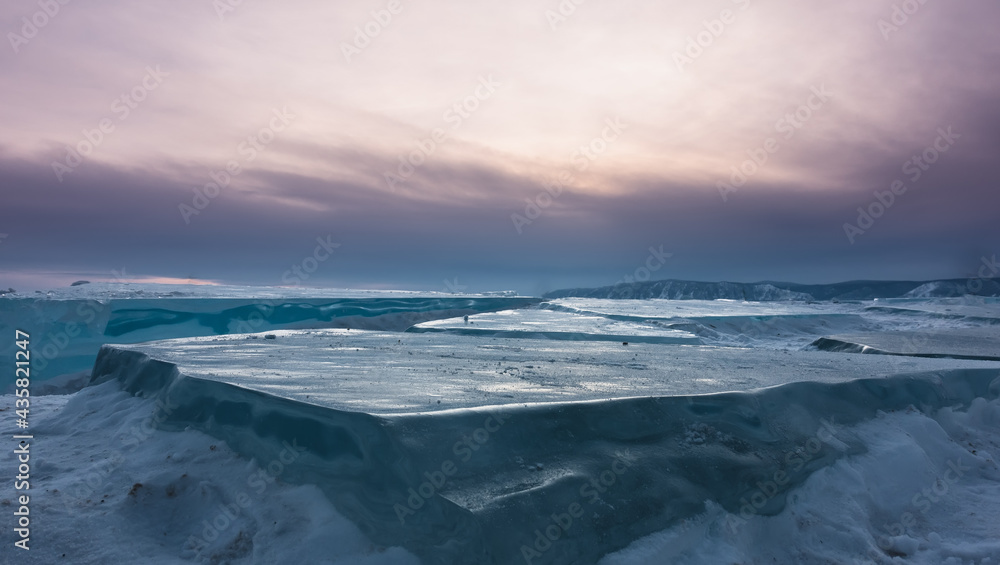Flat smooth ice floes on the surface of a frozen lake. Close-up. Pinkish evening sky, twilight. A mountain range on the horizon. Baikal