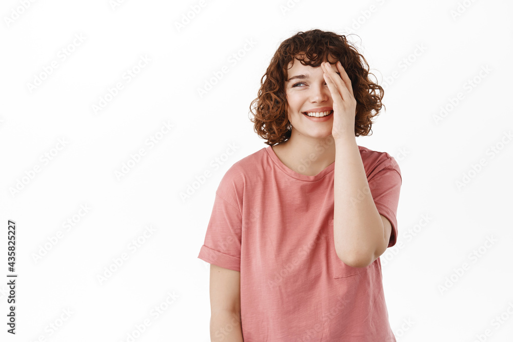 Beautiful young girl laughing, cover half of face and blushing cute, looking aside at promo beauty advertisement, standing in t-shirt against white background Beautiful young girl laughing, cover half of face and blushing cute, looking aside at promo beauty advertisement, standing in t-shirt against white background