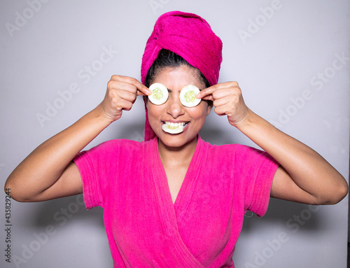 Beautiful Indian Lady in a pink bathrobe after bath giving beauty funny poses in front of the camera with body lotion and cucumber on her face © Eeshan