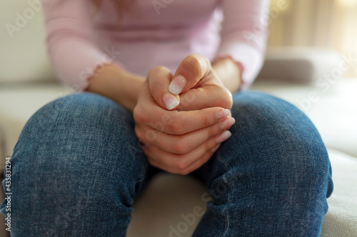 Cropped shot of a unrecognized young woman sitting on a sofa and feeling anxious. Close up image of woman feels nervous anxious or lonely put clenched hands on laps sitting on couch indoors.