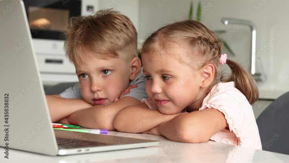two young children boy and girl sit at a table in the kitchen watching cartoons in a laptop and laughing. kids and gadgets. High quality 4k footage two young children boy and girl sit at a table in the kitchen watching cartoons in a laptop and laughing. kids and gadgets. High quality 4k footage