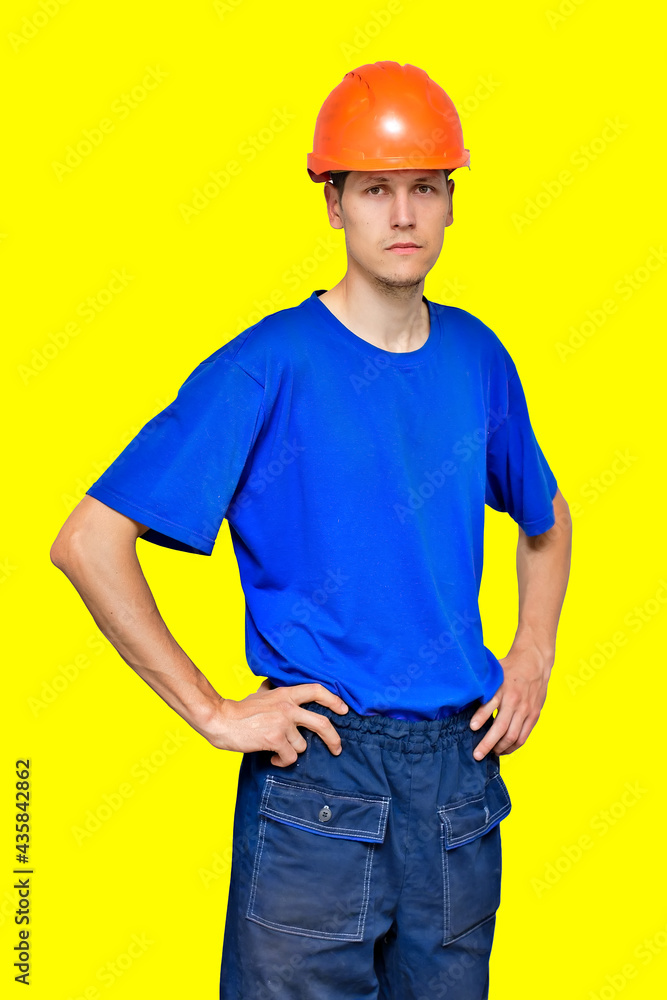 Portrait of a young thin worker in a construction helmet and a blue T-shirt. A builder or contractor is a neutral emotion. Isolated on a yellow background. Portrait of a young thin worker in a construction helmet and a blue T-shirt. A builder or contractor is a neutral emotion. Isolated on a yellow background.