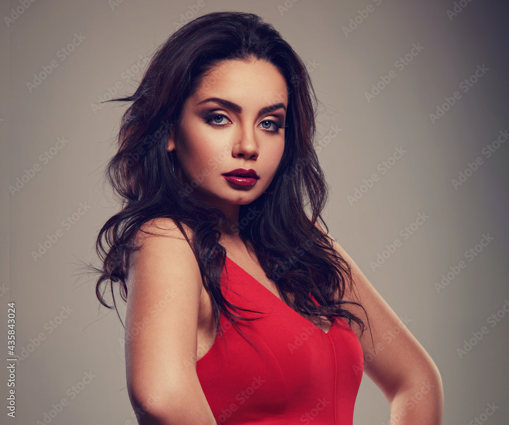 Fototapeta premium Beautiful makeup woman in red dress with long black curly volume hair style, burgundy lipstick with vamp look on grey background. Closeup
