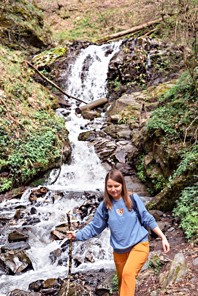 Young blond woman with stick in her hand going down stones along small waterfall in the mountain forest, mountain river, active lifestyle, digital detox, hiking alone