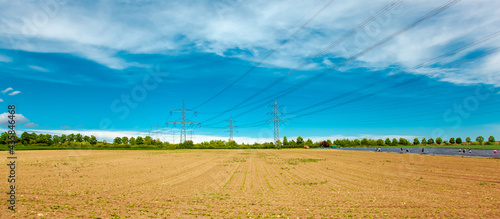 Agriculture field with high-tension pylons, power line transmission tower, High voltage towers against blue sky with clouds. Right hand side, Field of Asparagus covered with plastic foil with People