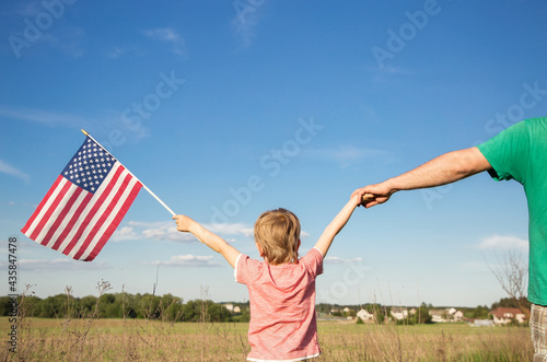 American flag in hand of child boy standing with his back against background of field. With other hand, hold on to his father's hand. Independence Day of United States of America. patriotic education