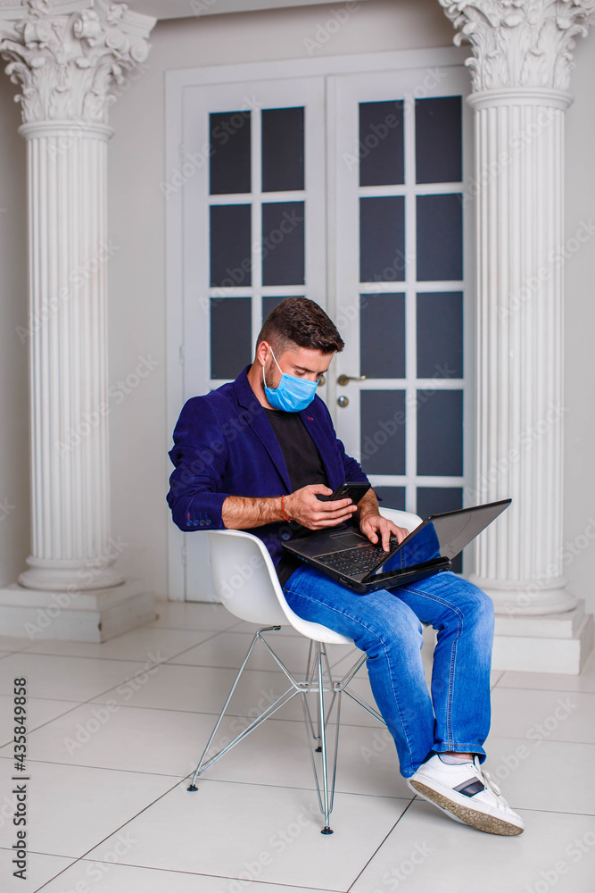A young man in a medical mask and a blue jacket sits on a chair and works on a laptop in a brightly lit room. internet office
