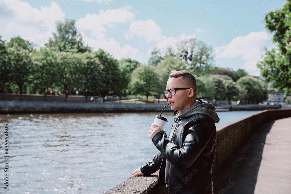 Young Caucasian man in eyeglasses, black jeans, leather jacket walks around city, drinks coffee and looks at river. Solo travel concept. Young Caucasian man in eyeglasses, black jeans, leather jacket walks around city, drinks coffee and looks at river. Solo travel concept.