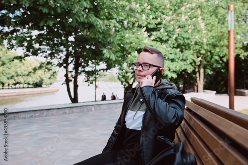Young caucasian man in black jeans, leather jacket sits on bench in city park and talks on smartphone. Rest in an urban environment, city life.