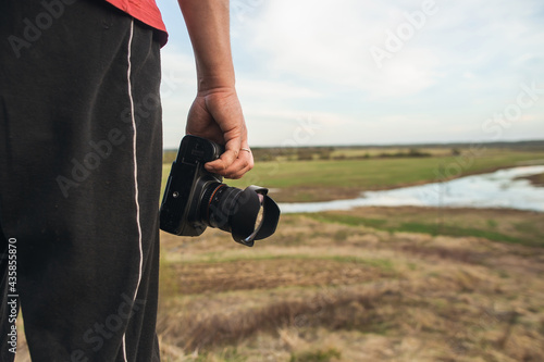 A male amateur photographer with a camera stands on a hill near a green meadow at sunset. Photorapher landscaper photo