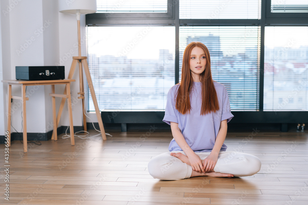 Portrait of young relaxed woman meditating sitting on floor in lotus position on background of window in bright office room. Attractive redhead lady performing relaxing breathing exercises at home.