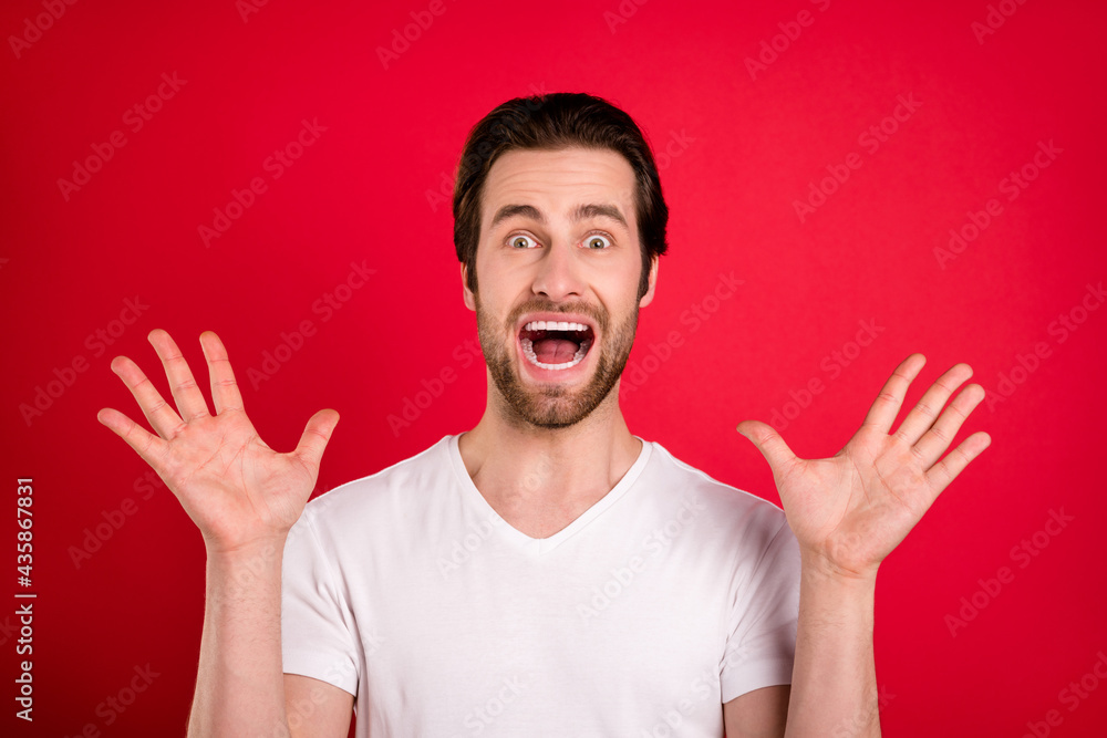 Photo of scared young brown hairdo man hands up wear white t-shirt isolated on vivid red color background Photo of scared young brown hairdo man hands up wear white t-shirt isolated on vivid red color background