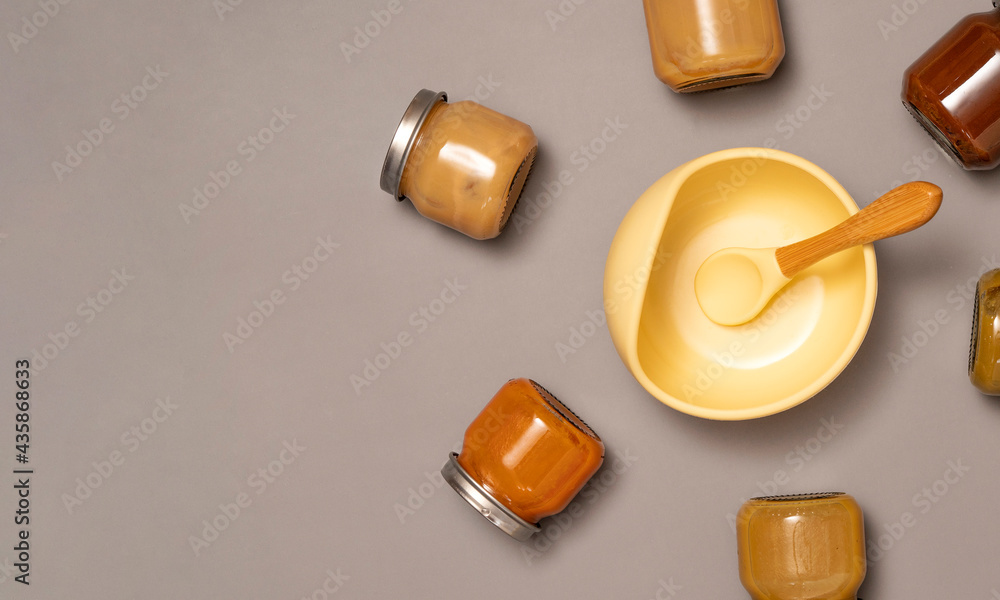 Glass jars with baby puree and children's tableware on grey background. First baby food. Lunch time concept. Glass jars with baby puree and children's tableware on grey background. First baby food. Lunch time concept.